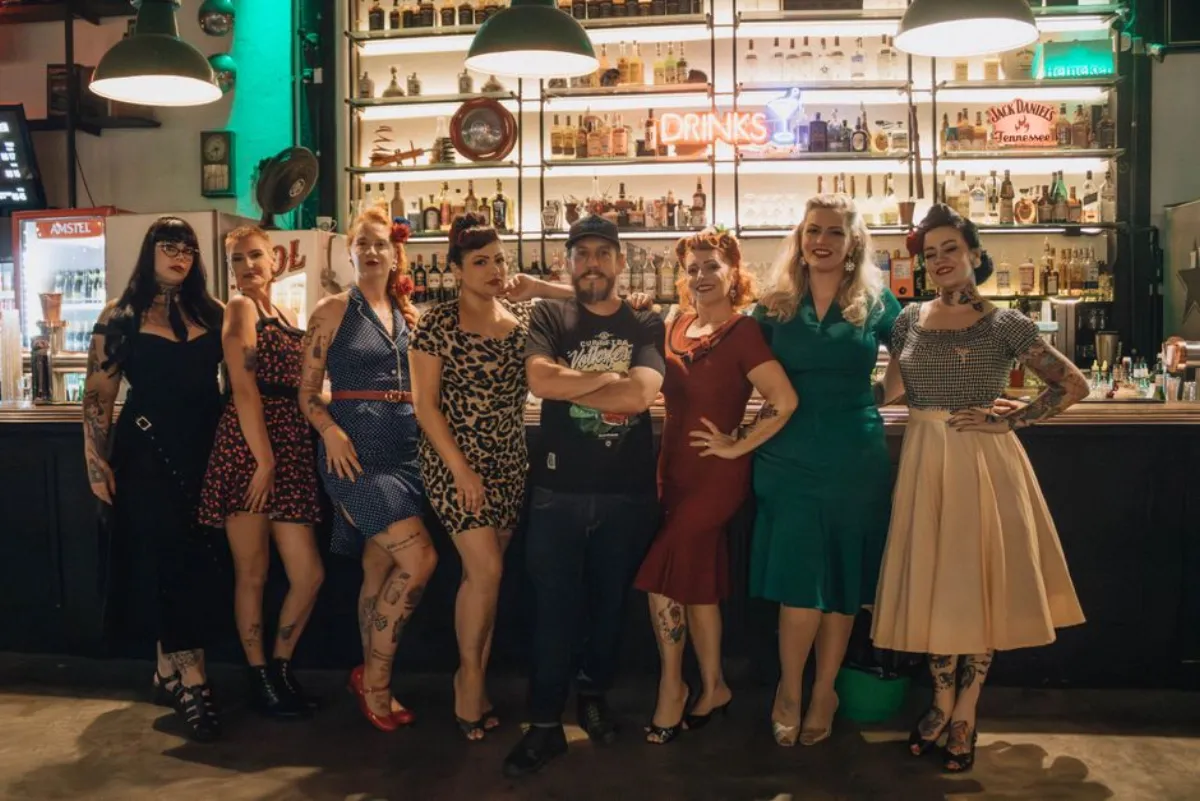 A lively group of adults in retro fashion poses at a stylish bar during the night.